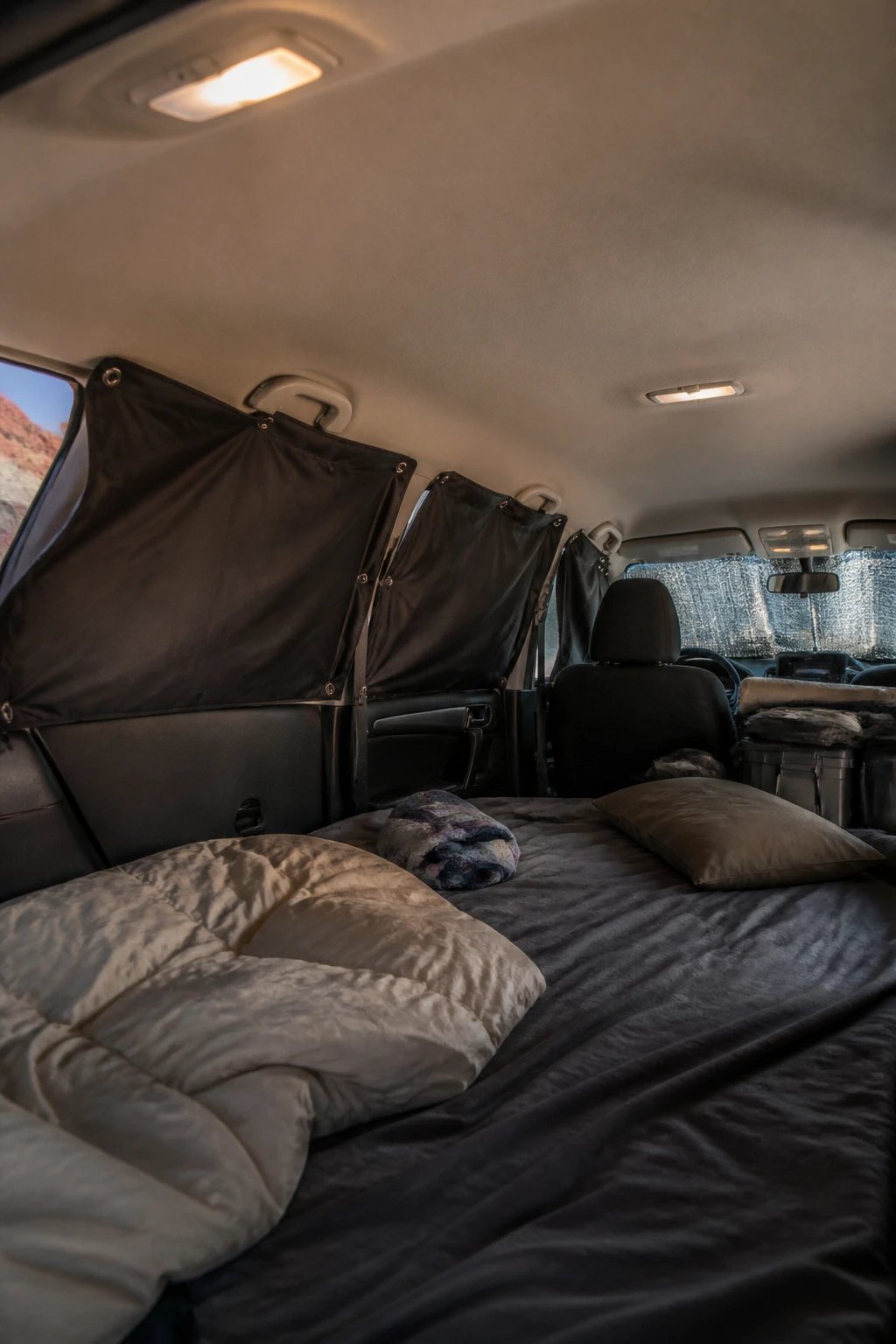A realistic photo of the interior of an SUV or hatchback with the rear seats folded down, showing a DIY privacy curtain system installed across the windows using magnetic clips holding black fabric curtains, with reflective sunshades visible in the windshield, soft ambient lighting inside creating a cozy private sleeping space, camping gear visible in the background, shot from inside the vehicle looking toward the covered windows, natural outdoor scenery barely visible through gaps in the curtains, professional automotive photography style with sharp focus and warm tones