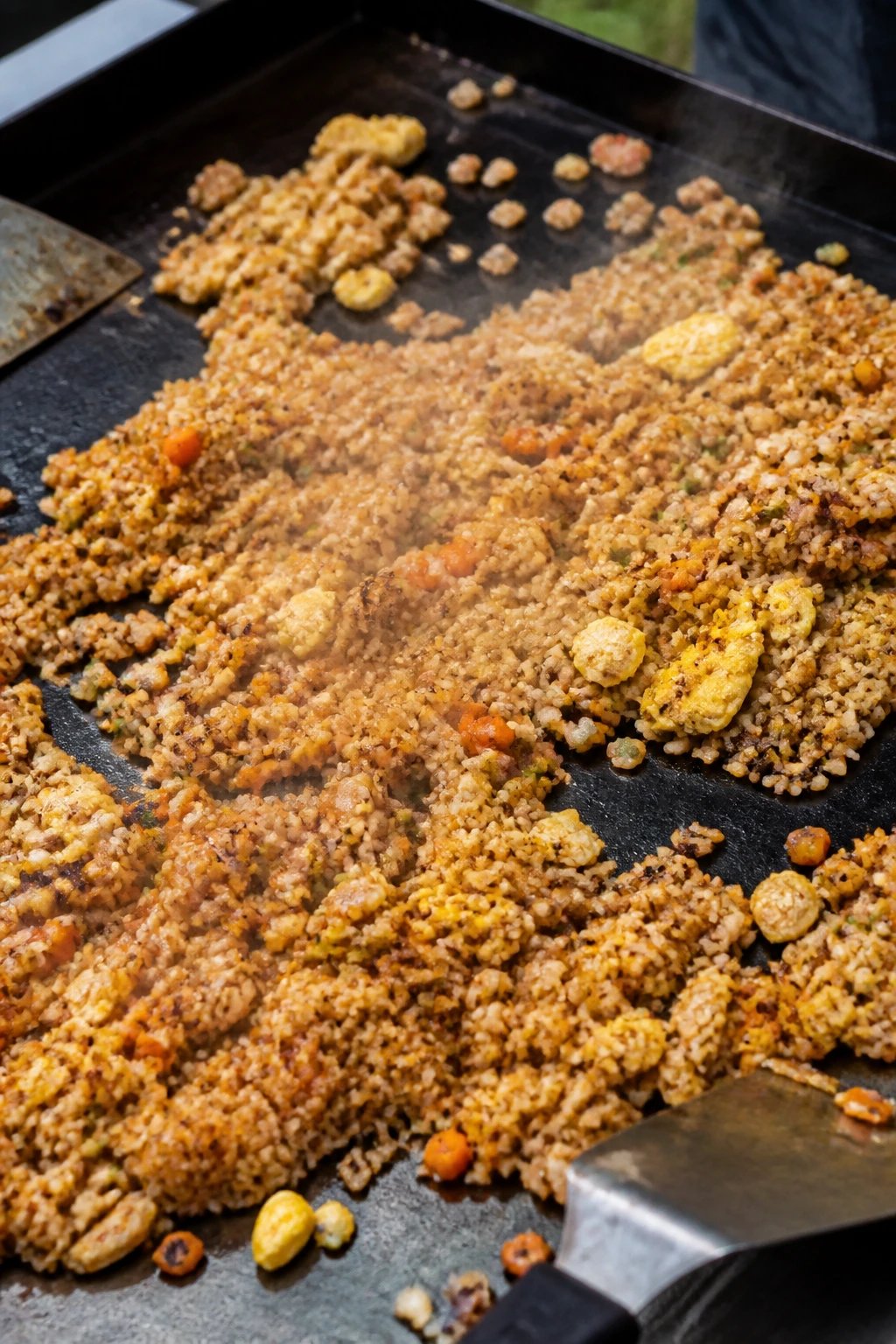 A high-quality overhead photograph of sizzling hibachi-style fried rice cooking on a flat top griddle, featuring golden-brown rice with visible char marks, colorful diced vegetables including bright orange carrots, scrambled egg pieces mixed throughout, steam rising from the hot surface, spatulas visible at the edge, the rice spread across the black cooking surface with some areas showing crispy caramelization, realistic outdoor cooking setting with natural daylight, professional food photography style with shallow depth of field