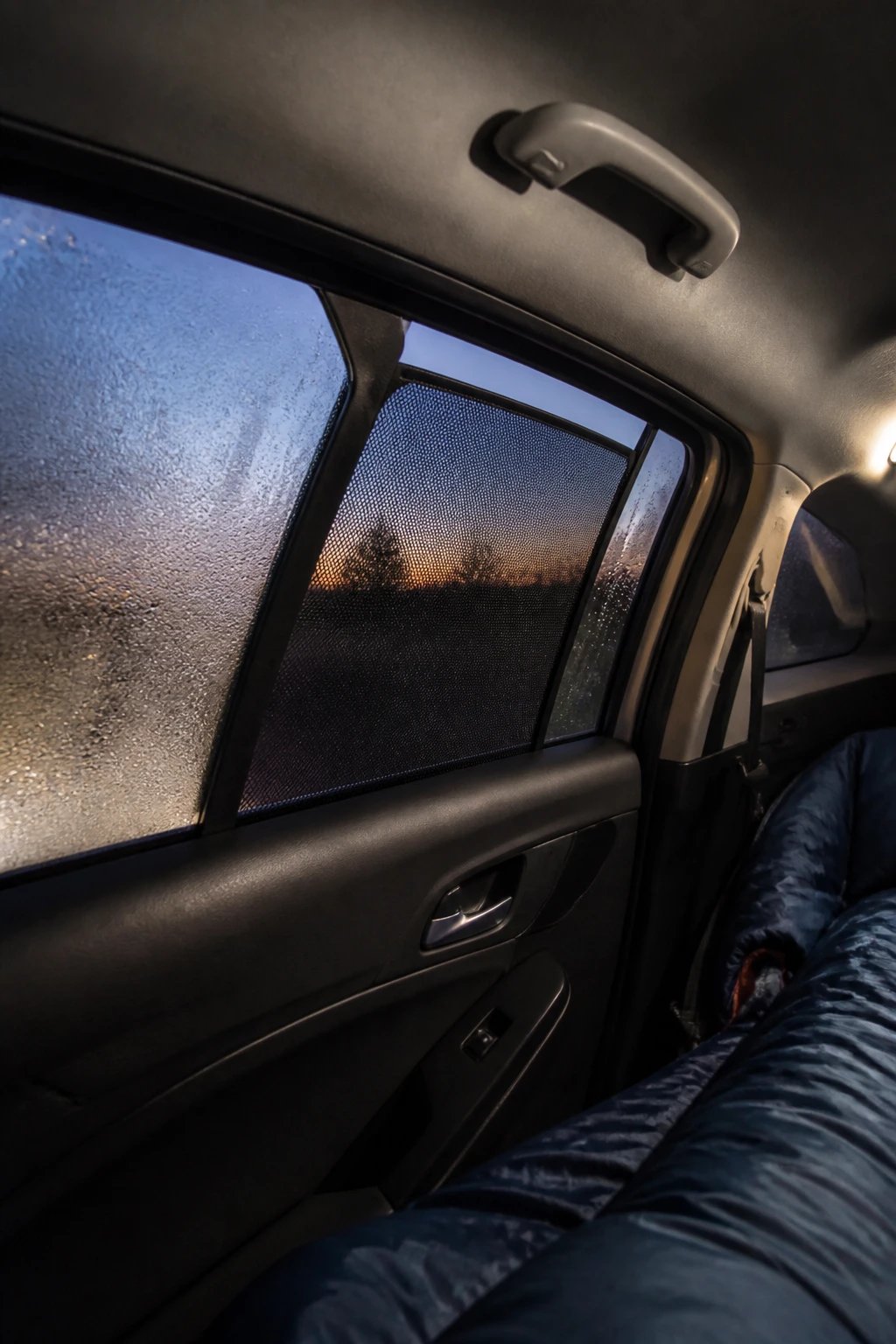 A realistic photograph showing the interior view of a compact car at dusk with windows slightly cracked open and mesh ventilation screens fitted into the window gaps, visible condensation droplets on parts of the glass contrasting with clear sections where air flows, a sleeping bag visible in the background, soft ambient lighting illuminating the ventilation setup, shot from inside the vehicle looking toward the window to showcase the anti-condensation mesh screen system in detail