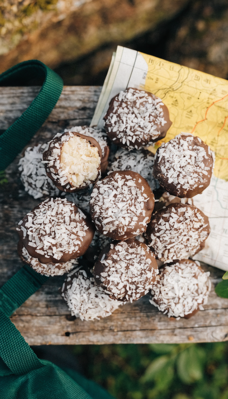 A high-quality overhead photograph of coconut-based energy bites (Packaroons) arranged on a rustic wooden surface with camping gear in soft focus in the background, showing the round, textured coconut macaroon-style snacks in dark chocolate coating, positioned next to a backpack strap and topographic map, with natural outdoor lighting creating subtle shadows, ultra-realistic product photography style, shallow depth of field, earthy tones with rich brown and cream colors, capturing the portable trail snack's texture and appetizing appearance in a wilderness camping setting.