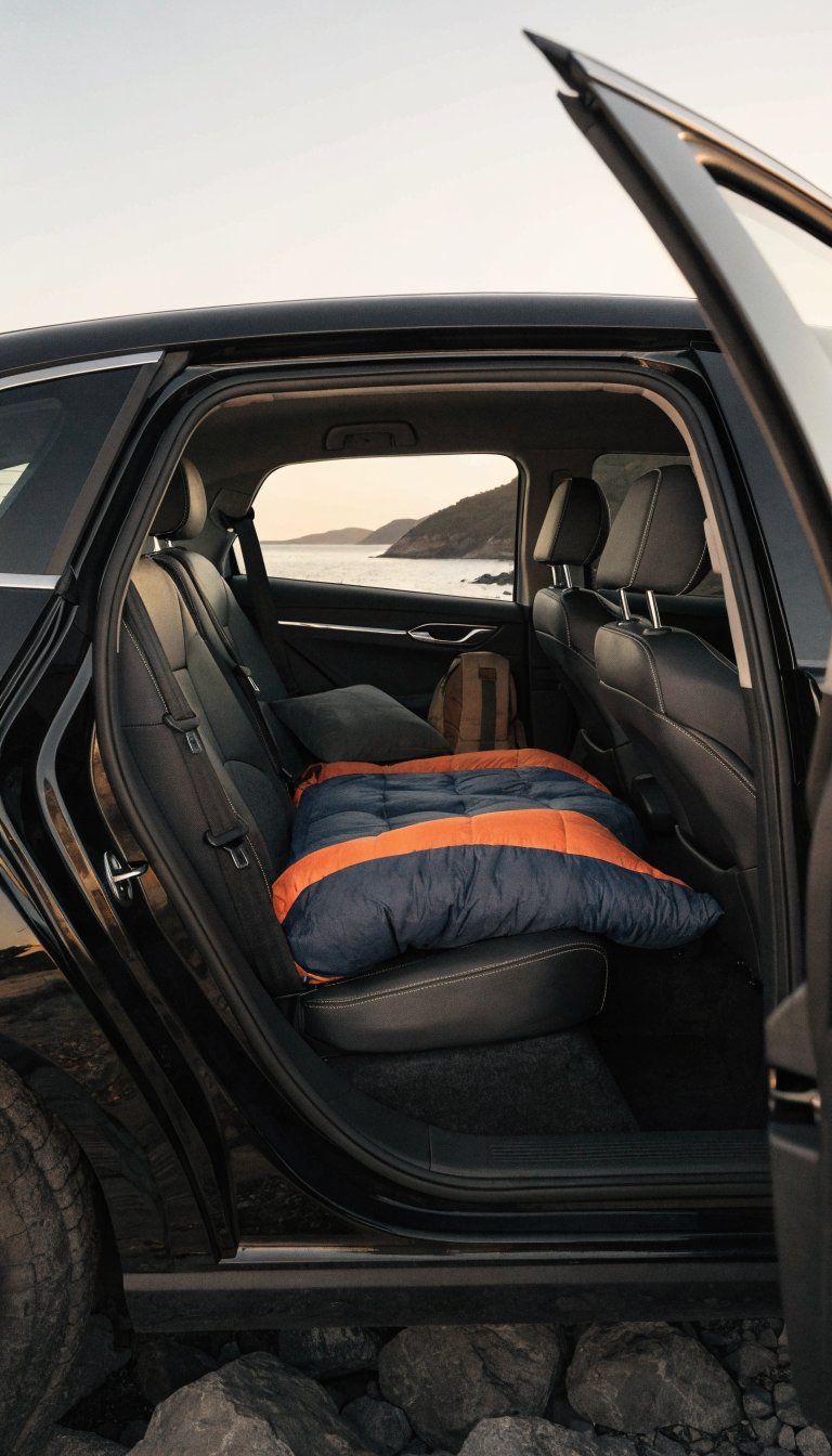 A realistic photograph taken from outside a sedan with the rear door open, showing the interior with both front seats folded completely forward and pushed to their maximum forward position, creating a flat sleeping platform in the back seat area. A sleeping bag is laid out diagonally across the expanded back seat space, demonstrating the camping setup. The image is shot during golden hour with soft natural lighting illuminating the car's interior, showing the practical conversion of a regular sedan into a sleeping space. The angle clearly displays how the folded front seats create additional room, with camping gear visible including a pillow and small backpack. High-quality automotive photography style with sharp focus on the interior layout.